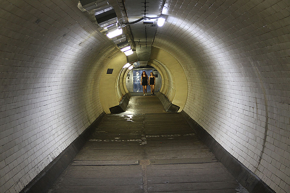 Děvčata v tunelu pod Temží pózují před skleněnou stěnou moderního výtahu vestavěného do šachty z roku 1906.