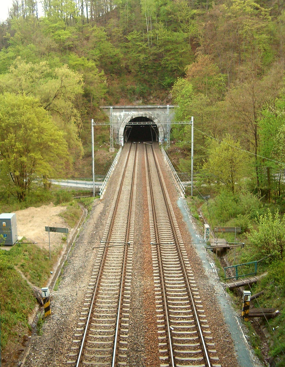 Pohled na Blanenský tunl č. 3 od severu, z jižního portálu <a href=/Tema/Blanensk%C3%BD%20tunel%20%C4%8D.%204>Blanenského tunelu č. 4</a>. Na plácku u reléové skříně vlevo stával přibližně do roku 2000 strážní domek, mezi tunely překonává trať řeku <a href=/Tema/Svitava>Svitavu</a>.